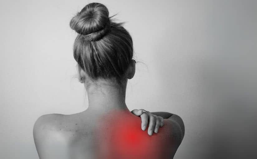 A woman grimaces, holding her shoulder, indicating discomfort and pain.