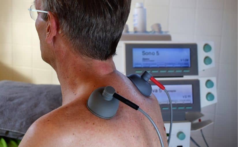 A man using a back massager while sitting in a hospital room, surrounded by medical equipment and a bed.