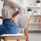 A woman sitting on a chair, visibly uncomfortable, holding her back in pain.