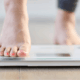 A person stands on a scale, balancing with her toes at the top, indicating she is checking her weight.