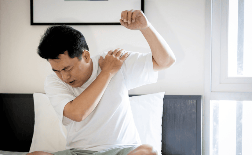 Man sitting on a bed, visibly distressed, holding his lower back due to pain.
