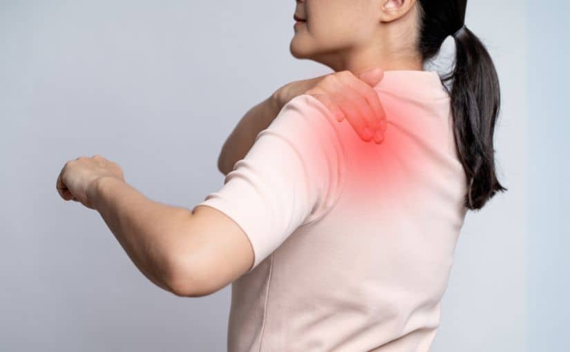 A woman grimaces, holding her shoulder in discomfort, indicating pain and distress in her expression