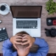 Overhead view of a person holding their head in frustration while working at a wooden desk with a laptop, camera, and slate board.