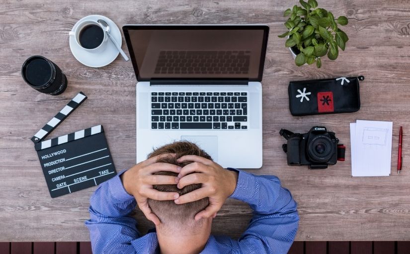 Overhead view of a person holding their head in frustration while working at a wooden desk with a laptop, camera, and slate board.