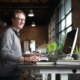 A smiling man with glasses standing and typing on a computer at a white height-adjustable standing desk in a modern office space.