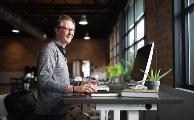 A smiling man with glasses standing and typing on a computer at a white height-adjustable standing desk in a modern office space.